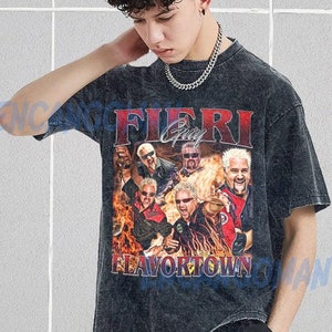 May include: A dark gray t-shirt featuring a graphic of Guy Fieri and the text "Flavortown" in red and white. The shirt has a vintage, distressed look and a crew neck.