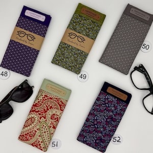 May include: An assortment of eyeglass cases in various colors and patterns, including purple, green, gray, red, and blue paisley. Each case has a "HANDMADE" label and a brown paper band with glasses illustration and "Handmade by Francie" text. Sunglasses and eyeglasses are also present.