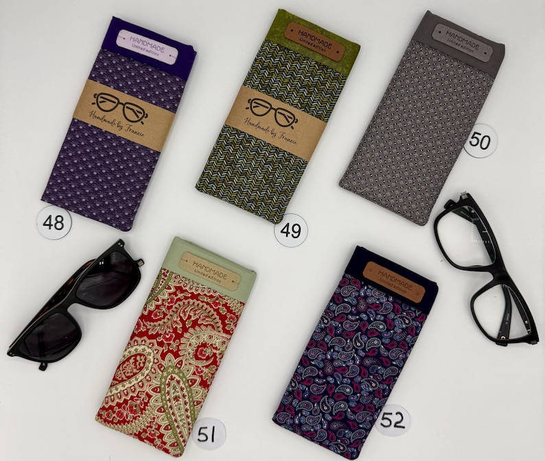 May include: An assortment of eyeglass cases in various colors and patterns, including purple, green, gray, red, and blue paisley. Each case has a "HANDMADE" label and a brown paper band with glasses illustration and "Handmade by Francie" text. Sunglasses and eyeglasses are also present.
