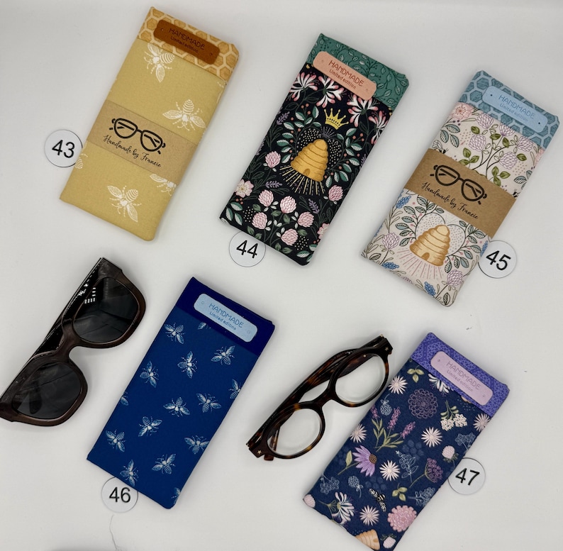 May include: A collection of handmade eyeglass cases in various patterns and colors. Designs include bees, floral, and insect motifs. Each case has a label that says "HANDMADE" and "Handmade by Francie".