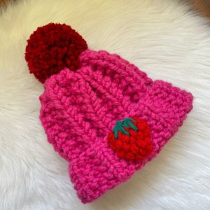 May include: A pink knitted beanie with a red pom pom and a red embroidered strawberry on the side.