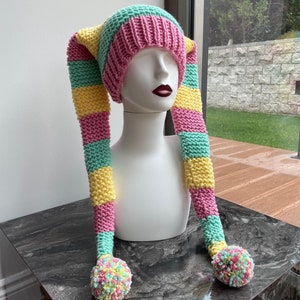 May include: A pink, yellow, and green striped knit hat with long, striped ear flaps and pom-poms on the ends.