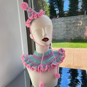 以下が含まれることがあります： ピンクとグリーンのクロシェ編み襟と帽子。襟はフリル付きで、帽子にはポンポンが付いています。