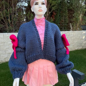 May include: A cropped blue knitted cardigan with large, puffy sleeves and pink bows on the cuffs. The cardigan is open in the front and has a V-neck.