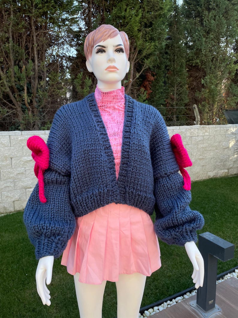 May include: A cropped blue knitted cardigan with large, puffy sleeves and pink bows on the cuffs. The cardigan is open in the front and has a V-neck.
