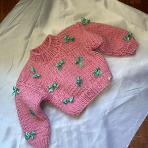 May include: A pink knitted sweater with green bows. The sweater has a crew neck and long sleeves.