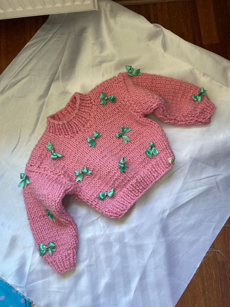 May include: A pink knitted sweater with green bows. The sweater has a crew neck and long sleeves.