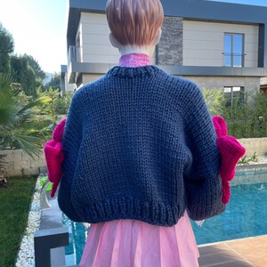 May include: A dark blue knitted sweater with large pink bows on the sleeves. The sweater is cropped and has a relaxed fit.