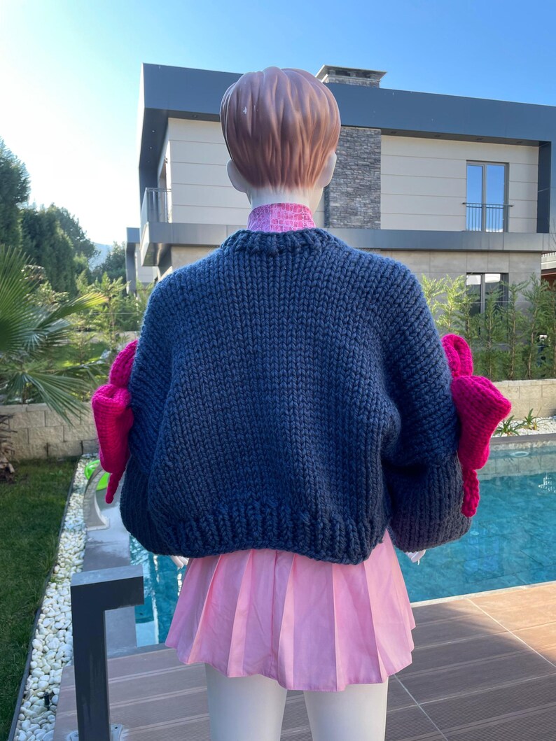 May include: A dark blue knitted sweater with large pink bows on the sleeves. The sweater is cropped and has a relaxed fit.