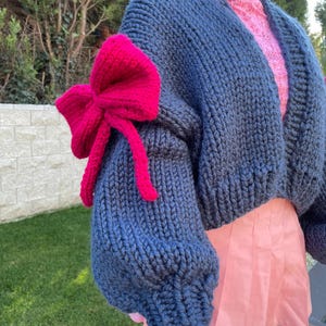 May include: A blue knitted cardigan with a large pink bow on the sleeve. The cardigan is cropped and has a relaxed fit.