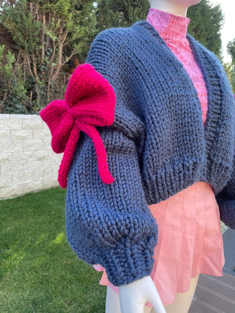 May include: A blue knitted cardigan with a large pink bow on the sleeve. The cardigan is cropped and has a relaxed fit.
