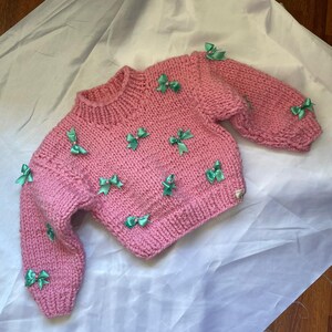 May include: A pink knitted sweater with green bows. The sweater has a crew neck and long sleeves.