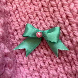 May include: A green satin bow with a pink pearl accent on a pink knitted background.