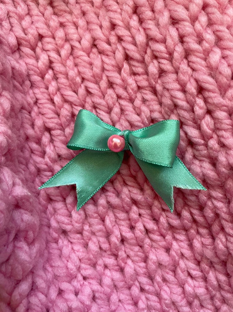 May include: A green satin bow with a pink pearl accent on a pink knitted background.