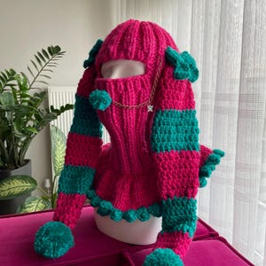 Può includere: Un passamontagna fatto a mano, dai colori vivaci rosa e verde acqua. Il cappello lavorato a maglia presenta una copertura per il viso, maniche lunghe a righe verde acqua e pompon verde acqua. Una catena dorata con un ciondolo a farfalla aggiunge un tocco decorativo.