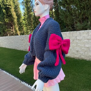 May include: A blue knitted cardigan with pink bow details on the sleeves. The cardigan is cropped and has a relaxed fit.
