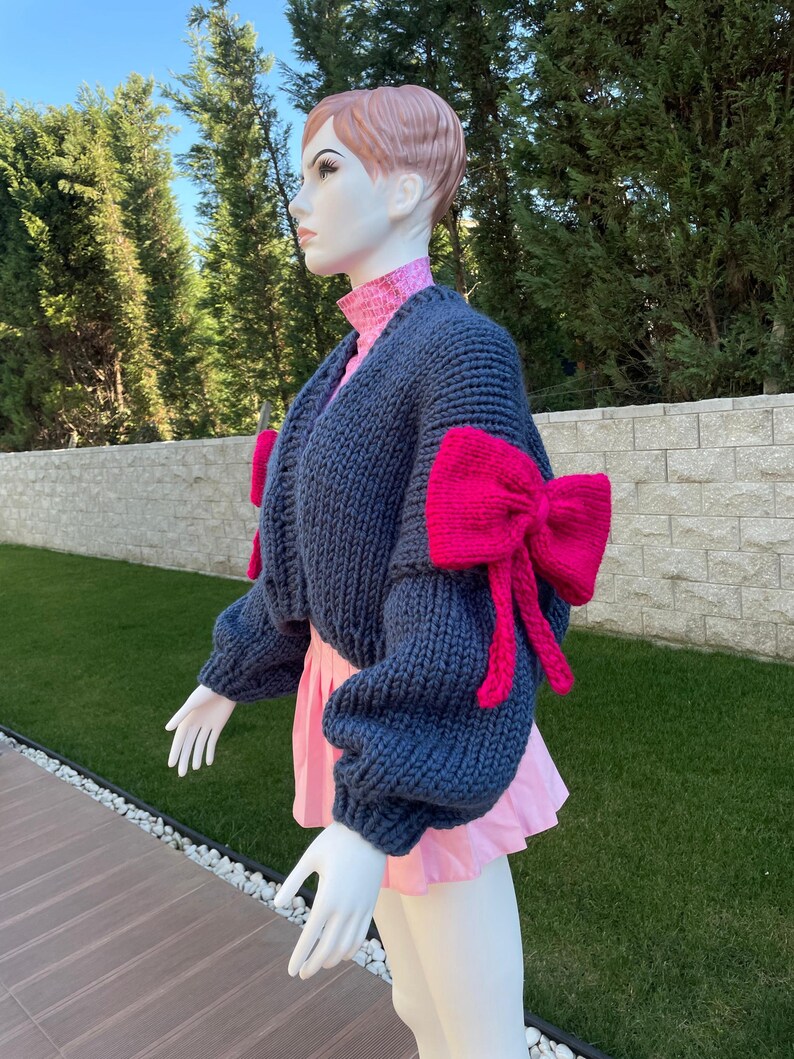May include: A blue knitted cardigan with pink bow details on the sleeves. The cardigan is cropped and has a relaxed fit.