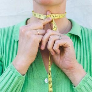 以下が含まれることがあります： 人が黄色のメジャーで首を測っています。メジャーにはインチで数字が記されています。その人は緑色のシャツを着ています。