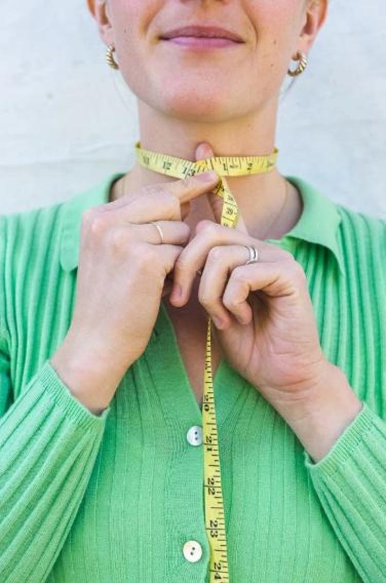 以下が含まれることがあります： 人が黄色のメジャーで首を測っています。メジャーにはインチで数字が記されています。その人は緑色のシャツを着ています。