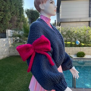May include: A navy blue knitted sweater with a large pink bow on one sleeve. The sweater has a relaxed fit and is made of a soft, chunky yarn.