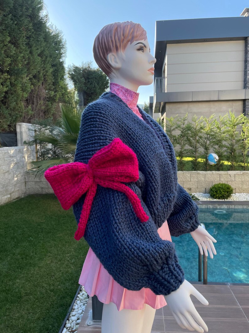 May include: A navy blue knitted sweater with a large pink bow on one sleeve. The sweater has a relaxed fit and is made of a soft, chunky yarn.