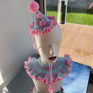 以下が含まれることがあります： 帽子と襟のセット。ピンクとグリーンの編み物で、帽子のてっぺんにポンポンがついています。