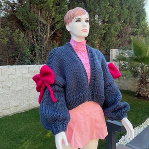 May include: A cropped blue knitted cardigan with large pink bows on the sleeves. The cardigan is worn over a pink top and a pink skirt.
