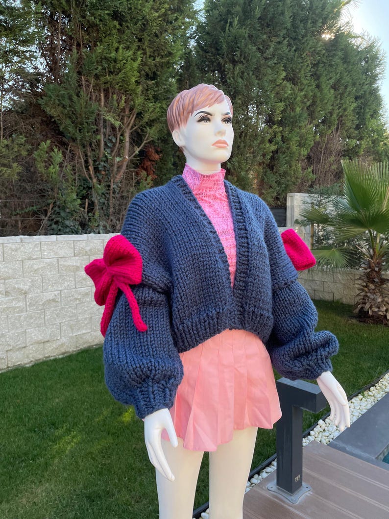 May include: A cropped blue knitted cardigan with large pink bows on the sleeves. The cardigan is worn over a pink top and a pink skirt.