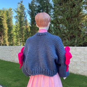 May include: A blue knitted sweater with pink bows on the sleeves. The sweater is cropped and has a loose fit.
