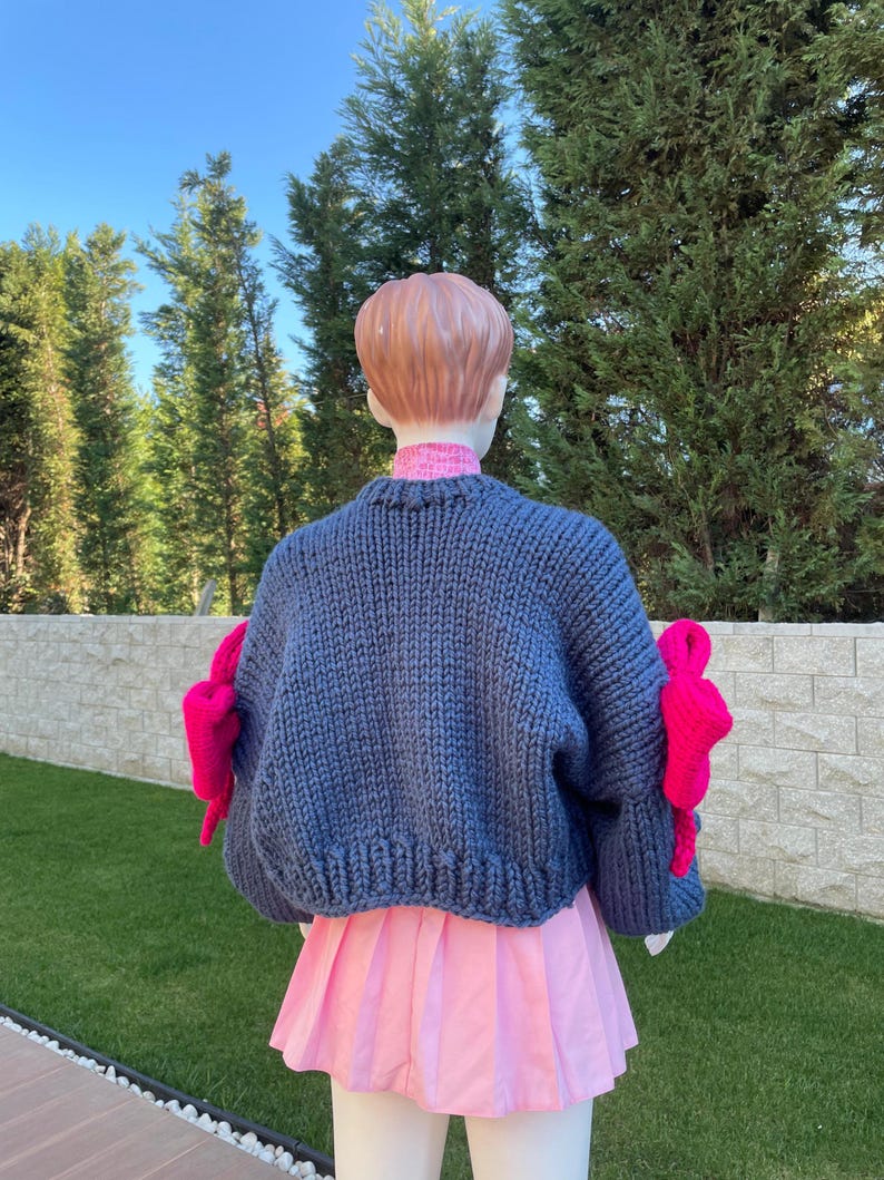 May include: A blue knitted sweater with pink bows on the sleeves. The sweater is cropped and has a loose fit.
