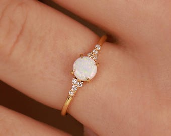 Puede incluir: Un anillo de oro con una piedra de ópalo blanca redonda central, flanqueada por tres pequeñas piedras transparentes a cada lado. La fina banda de oro pulido sujeta las piedras en un engaste de garras, creando un aspecto delicado y refinado.