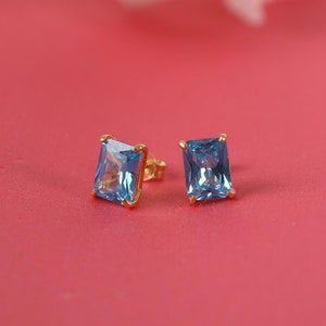 May include: A pair of gold stud earrings with blue square-shaped gemstones. The earrings are on a pink surface.