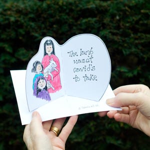 May include: A white pop-up card held by hands, featuring a watercolor illustration of a woman in a pink dress holding a lamb, with two children. The text reads "The lamb wasn't David's to take."