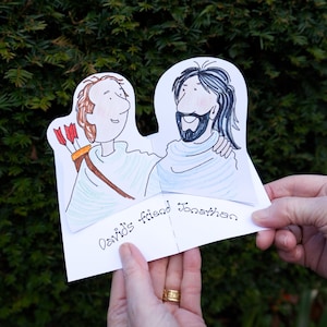 May include: A hand-drawn card featuring two cartoon figures embracing. One figure has a bow and arrows, the other has a beard. The text "David's friend Jonathan" is written on the card. The card is held in front of a green leafy background.