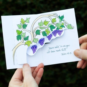 May include: A white card with a drawing of a vine with purple grapes. The card is folded in half and the text "Jesus said, 'In me you will bear much fruit.' John 15:5" is visible on the right side of the card.