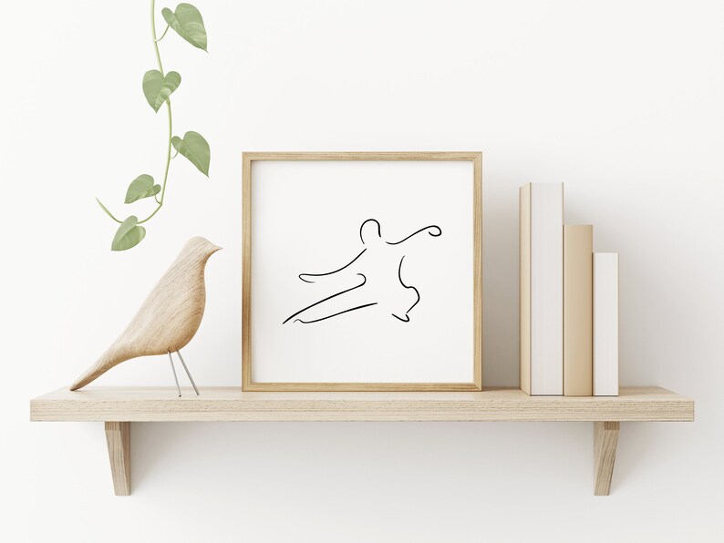 May include: A framed black and white line drawing of a person in a martial arts pose, on a wooden shelf with a wooden bird figurine and a potted plant.