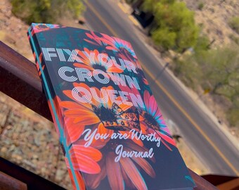 May include: A black journal with a floral design featuring orange, pink, and blue flowers. The text "Fix Your Crown Queen You Are Worthy Journal" is printed on the front cover.