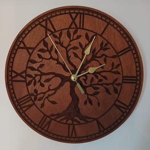 May include: A circular wooden wall clock with a tree of life motif. The clock face displays Roman numerals and gold-coloured hands. The tree design is etched into the wood, creating a detailed decorative element. The clock is a warm brown colour.