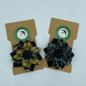 May include: Two fabric flower brooches with black centres and plaid fabric in yellow, green, and black. The brooches are on brown cards with the text "Swallow's Nest Design".