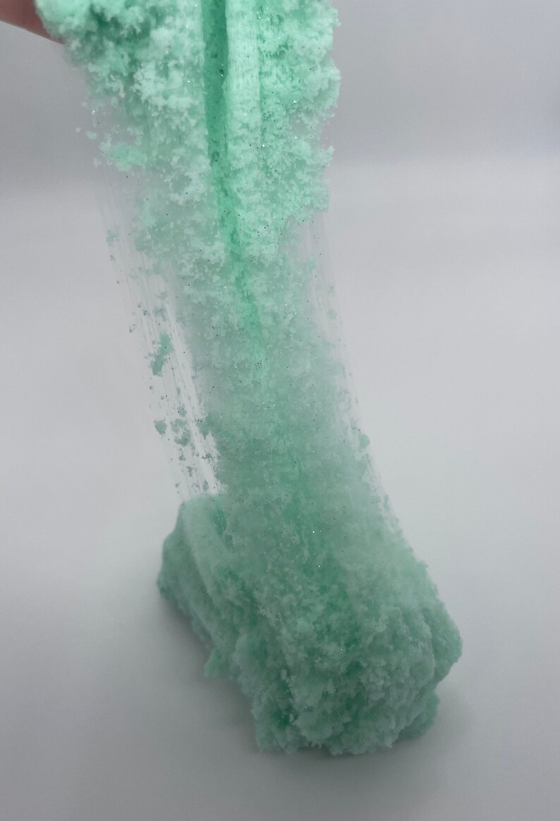 May include: A light green slime with a glittery texture. The slime is being stretched and pulled, revealing its stringy consistency.