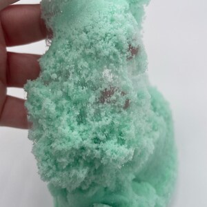 May include: Close-up of a light green slime with a sparkly, glittery texture. The slime is being held in a hand and is very thick and gooey.
