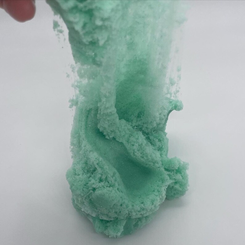 May include: A close-up of a light green slime with a grainy texture. The slime is being pulled up and away from a white surface.