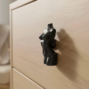 May include: A black, female torso-shaped cabinet knob. The knob is mounted on a light-coloured wooden cabinet. The design is a minimalist, modern take on a classic form, adding a unique touch to furniture.