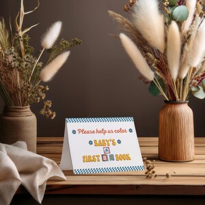 May include: A white card with blue and yellow checkered border. The text on the card reads "Please help us color * Baby's * A B Book * First *"