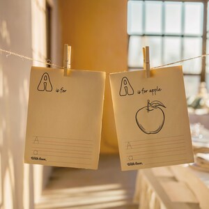 May include: Two blank cards with clothespins hanging from a string. The cards are beige and have the letter "A" written on them. One card says "A is for apple" and has a drawing of an apple. The other card says "A is for" and has a blank line for writing.