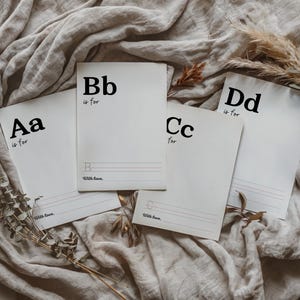 May include: Four white cards with black lettering. Each card features a letter of the alphabet, 'Aa', 'Bb', 'Cc', and 'Dd', with the phrase 'is for' written below. The cards are on a beige fabric background.