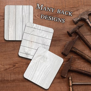 May include: Three white wood coasters with a wood grain pattern. The coasters are on a brown wood surface. The text "MANY BACK DESIGNS" is written in black letters on the surface.