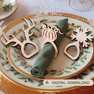 Può includere: Tre portatovaglioli in legno chiaro con disegni tagliati al laser a forma di corna di cervo, zucca e fiocco di neve. Un anello trattiene un tovagliolo di stoffa verde oliva arrotolato. L'immagine include il testo DIGITAL DOWNLOAD.
