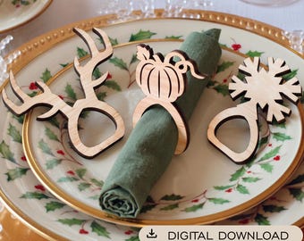 Puede incluir: Tres servilleteros de madera clara con diseños cortados a láser de una cornamenta de ciervo, una calabaza y un copo de nieve. Un anillo sostiene una servilleta de tela verde oliva enrollada. Incluye el texto DIGITAL DOWNLOAD.