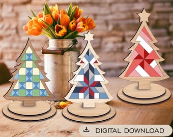 May include: Three wooden Christmas tree ornaments with geometric quilt-style patterns. The left tree features green and blue squares, the center tree has blue, red, and white triangles, and the right tree displays red and pink segments.